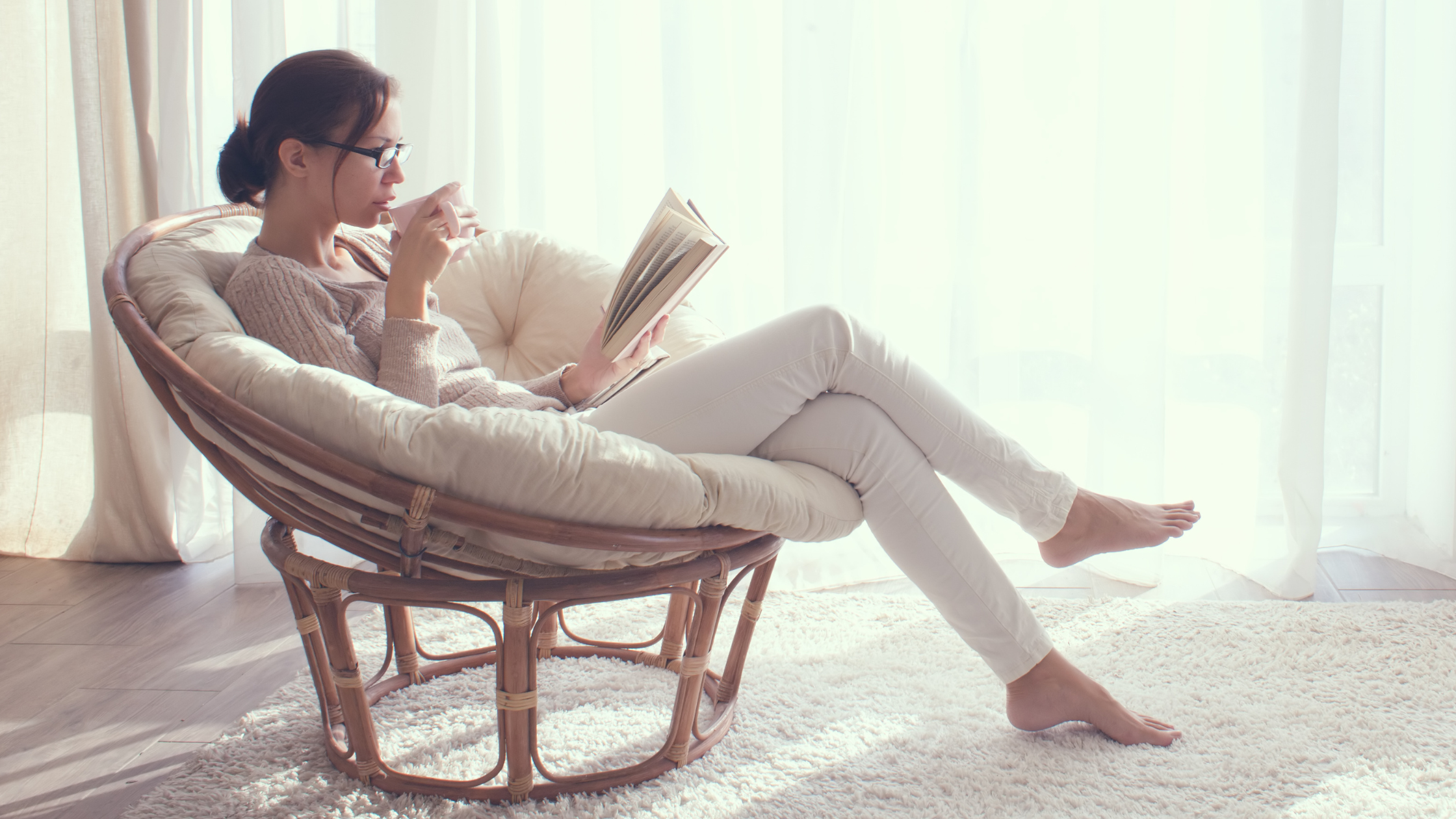 Frau in sonnendurchflutetem Raum sitzt in bequemen Korbsessel, enstpannt, Buch lesen, Getränk trinken Pragmatische Energetik